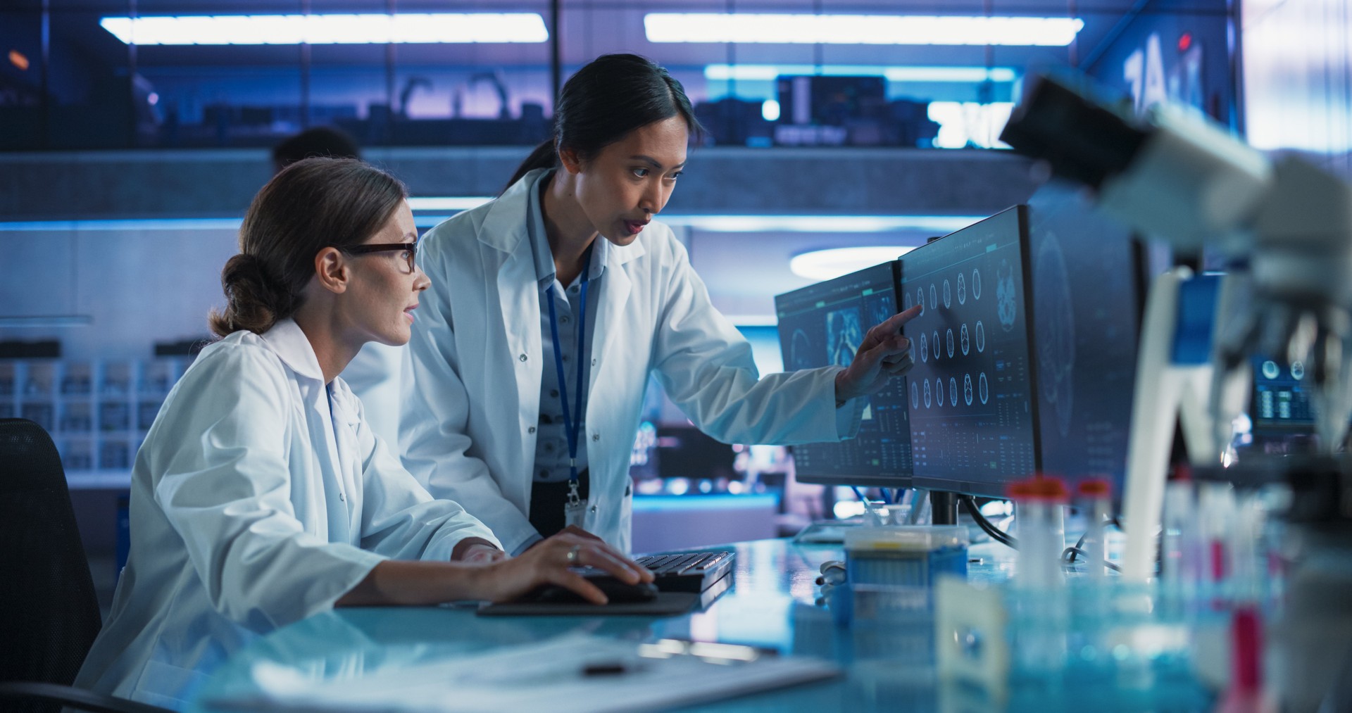 Asian And Caucasian Female Doctors Talking In Medical Research Center And Using Desktop Computer To Analyze MRI Scans Of Brain. Surgeon And Neuroscientist Discussing Solutions For Treating Cancer.
